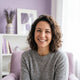 A smiling woman in her late 30s looks at the camera with a warm, contented expression in a bright home setting.